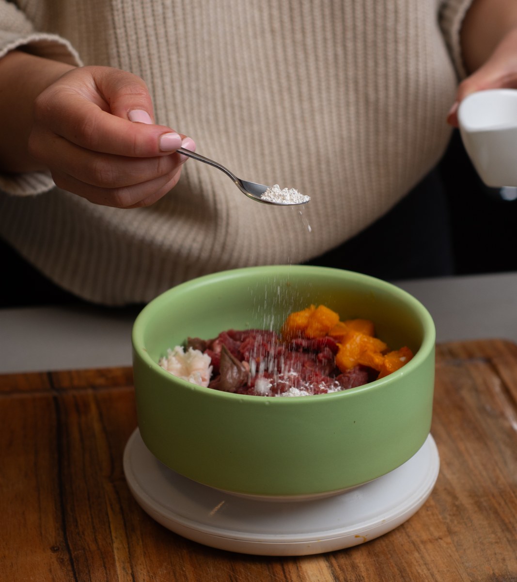 A spoonful of supplement powder being sprinkled over a green bowl of fresh dog food with beef, liver and pumpkin