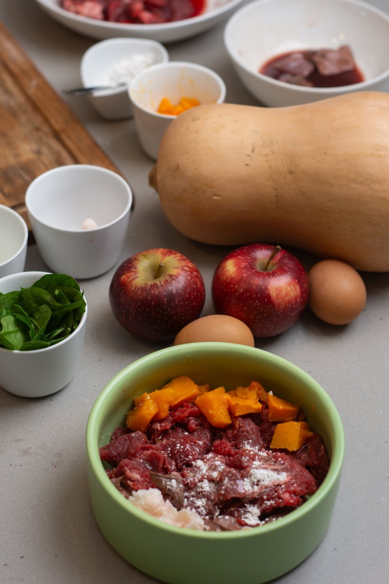 A French Bulldog meal in a green bowl: raw beef and liver topped with cubes of cooked butternut pumpkin, with a whole butternut, apples and eggs on the bench behind