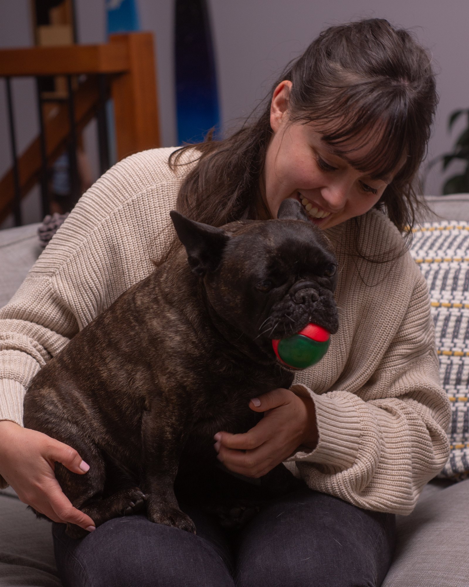 Alessa Grimm on the couch cuddling her brindle French Bulldog Audrey, who is holding her favourite ball
